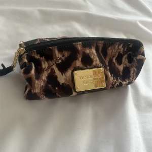 Leopardmönstrad necessär från Victoria's Secret - Snygg leopardmönstrad necessär från Victoria's Secret med gulddetaljer och dragkedja. Perfekt för att hålla ordning på dina småsaker. Den har en röd insida som ger en fin kontrast till det djuriska mönstret.