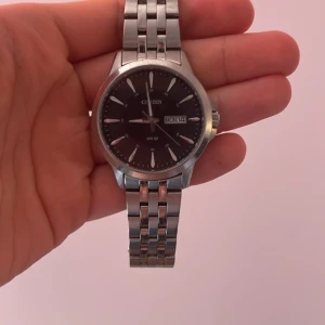 Silverfärgad klocka från Citizen - Stilren klocka från Citizen med silverfärgad länkarmband och svart urtavla. Klockan har dag- och datumvisning samt tydliga timmarkeringar. Perfekt för den som söker en klassisk och tidlös accessoar.