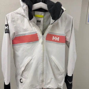 Vit jacka från Helly Hansen - Säljer en vit jacka från Helly Hansen med dragkedja och två fickor framtill. Jackan har svarta detaljer och en rosa rand över bröstet. Perfekt för utomhusaktiviteter nu i vår och sommar då den är en vindjacka! Nytvättad, fläckarna går säkert bort om man orkar lägga tid på det!