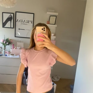 Ljusrosa topp med volangärmar från Gina Tricot - Säljer en söt ljusrosa topp från Gina Tricot med volangdetaljer på ärmarna. Toppen har en rund halsringning och är perfekt för en trendig look. Passar bra till både jeans och kjol. 🌸