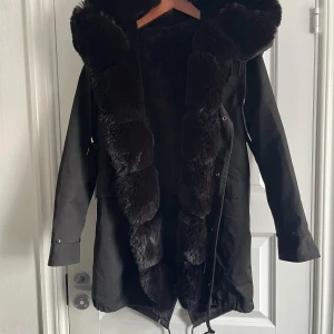 Svart parkajacka med pälsdetaljer - Snygg svart parkajacka med pälsdetaljer på huva och ärmslut. Jackan har används ett fåtal gånger och är i nästintill i nytt skick. Pälsen inuti går att ta bort med en dragkedja ifall man vill använda den höst, vinter eller vår. Pälsjacka eller skaljacka.