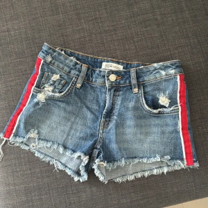 Lågmidjade jeansshorts - Lågmidjade snygga jeansshorts med slitna detaljer och fransar. De har röda och vita ränder längs sidorna som ger en sportig look. Perfekta för en avslappnad stil. 