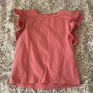 Rosa topp med volangärmar från 157 - Säljer en söt rosa topp från 157 med volangdetaljer på ärmarna. Toppen är tillverkad i en mjuk bomullsblandning och har en rund halsringning. Perfekt för en avslappnad stil.