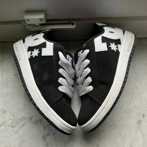 Svarta sneakers från DC Shoes - Snygga svarta sneakers från DC Shoes med vita detaljer och snörning. Skorna har en robust sula och är perfekta för en casual look. De har en vit stjärndetalj på sidan och en bekväm passform. Nästan helt nya