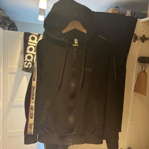 Moschino set, både byxor och hoodie nypris=5300kr - Snyggt svart sett från Moschino med vita och röda sidoremsor där märkesnamnet upprepas. Byxorna har en bakficka med Moschino-logga och en bekväm passform. Jag söker både tröjan och byxorna för de priset som står på annonsen. De är äkta och är köpta från U&M på VÄLA som ett sett för 5300kr
