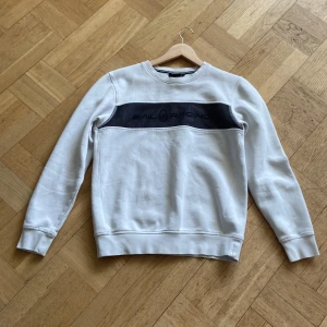 Vit tröja från Sail Racing - Snygg vit tröja från Sail Racing med ett svart band över bröstet där loggan är tryckt. Tröjan har långa ärmar och en rund halsringning.