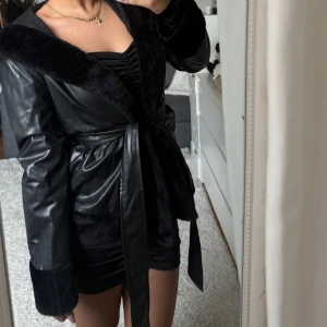 Svart skinnjacka med pälsdetaljer - Snygg svart skinnjacka med mjuka pälsdetaljer och knytbälte i midjan. Jackan har långa ärmar och en elegant design som passar perfekt för kyligare dagar.