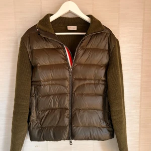 Olivgrön cardigan från Moncler - Snygg olivgrön jacka från Moncler med quiltad framsida och stickade ärmar. Jackan har en dragkedja framtill och en hög krage för extra värme. Perfekt för kyligare dagar med sin stilrena design och bekväma passform.