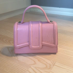 Rosa handväska med kedja - Snygg rosa handväska med krokodilmönster och en stilren kedja som axelrem. Perfekt för att ge en färgklick till din outfit. Väskan har ett praktiskt handtag och en elegant design.