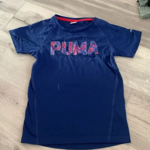 Blå t-shirt från Puma - Säljer en blå t-shirt från Puma med korta ärmar och ett stort Puma-tryck i rött på bröstet. Perfekt för träning eller vardagsbruk. Materialet är lätt och andas bra. Passar dig som gillar sportig stil!* Barnsize 