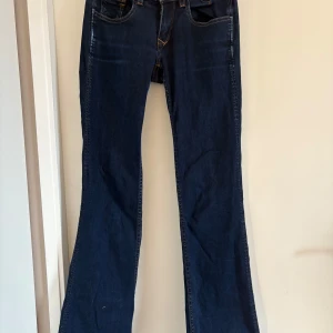 Mörkblå bootcut jeans från Hilfiger Denim - Snygga mörkblå bootcut jeans från Hilfiger Denim. De har en klassisk femficksdesign med guldfärgade knappar och kontrastsömmar. Mått: midja: 66 cm, innerben: 80 cm.