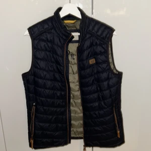 Svart/mörkblå väst från Camel Active - Snygg svart quiltad väst från Camel Active med dragkedja och bruna detaljer. Perfekt för lager-på-lager under kyliga dagar. Västen har en stilren design med en liten logotyp på bröstet. Storlek S funkier nog också för M