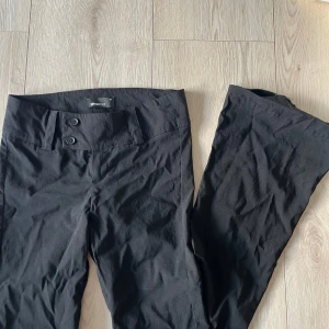 Svarta byxor från Gina Tricot - Snygga svarta byxor från Gina Tricot med låg midja och två knappar framtill. Perfekta för en stilren look. Byxorna har en rak passform och är tillverkade i ett bekvämt material.🩷🩷🩷