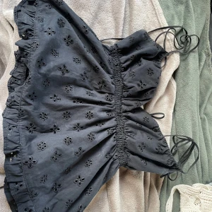 Babydoll topp - Snygg svart axelbandslös topp med elastisk smockdetalj och broderade blommor. Toppen har volangdetaljer och knytband för en justerbar passform. Perfekt för en somrig look!