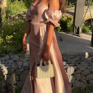 Champagnefärgad axelbandslös klänning - Elegant champagnefärgad axelbandslös klänning med puffiga ärmar och en hög slits. Klänningen har en vacker omlott-design och är dekorerad med pärldetaljer i midjan.