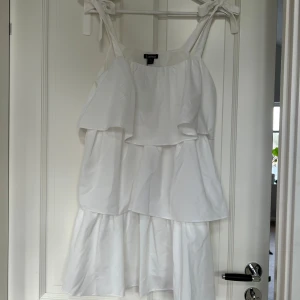Vit klänning från Boohoo - Säljer en söt vit klänning från Boohoo med volanger och knytbara axelband. Perfekt för sommarens alla tillfällen. Klänningen har en luftig passform och är tillverkad i ett lätt material. 
