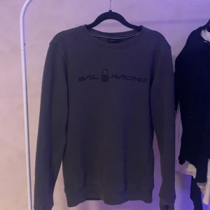 Grå tröja från Sail Racing - Säljer en stilren grå tröja från Sail Racing med deras logga tryckt på framsidan. Tröjan har långa ärmar och en rund halsringning. Perfekt för en avslappnad stil.
