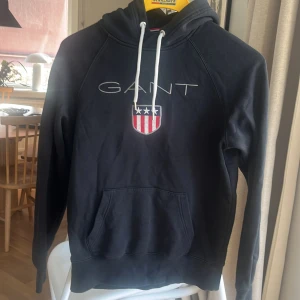Svart hoodie från GANT - Säljer en svart hoodie från GANT med vit dragsko och ett broderat emblem på bröstet. Perfekt för en avslappnad stil. Hoodien har en klassisk känguruficka framtill.