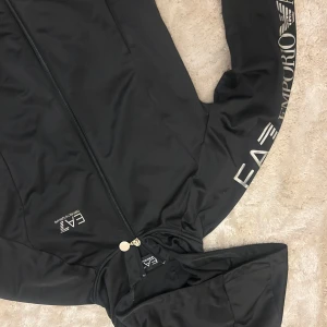 Svart hoodiejacka från Emporio Armani EA7 - Säljer en svart hoodiejacka från Emporio Armani EA7 med dragkedja framtill och tryckt logga längs ena ärmen. Jackan har huva och två fickor framtill. Perfekt för en sportig och stilren look.