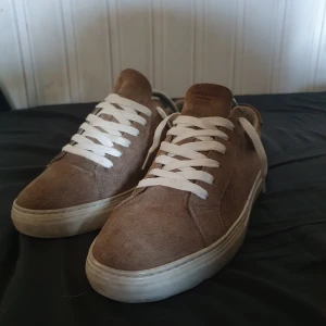 Bruna sneakers från Vagabond - Säljer ett par bruna sneakers från Vagabond i mocka med vita snören och platt sula. Skorna har en klassisk rund tå och passar perfekt till en avslappnad stil. Storlek 43.