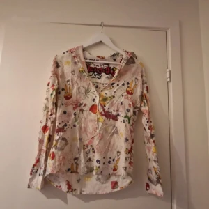 Färgglad långärmad skjorta med konstnärligt mönster - Säljer en unik och färgglad långärmad skjorta med v-ringning och knappar. Skjortan har ett konstnärligt, abstrakt mönster i flera färger som ger en kreativ vibe. Perfekt för dig som vill sticka ut med din stil.