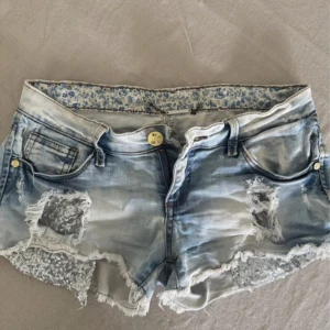 Blå jeansshorts med paljettdetaljer från Seventy Chicks - Säljer ett par ljusblå jeansshorts med slitna kanter och silvriga paljettdetaljer på framsidan. Shortsen har fem fickor, låg midja och råa benslut. Perfekta för en trendig sommarlook.
