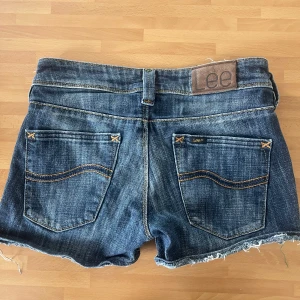 Mörkblå jeansshorts från Lee - Säljer ett par mörkblå jeansshorts från Lee med klassiska fem fickor och råa, fransiga benslut. Shortsen har normal passform och låg midja. Perfekta för varma dagar och enkel att matcha med olika toppar.