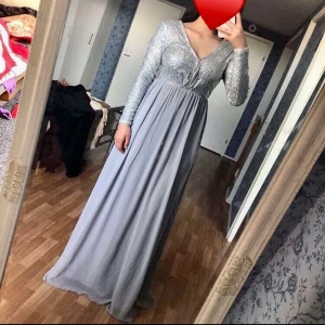 Silverglittrig långklänning med v-ringning - Säljer en elegant långklänning med glittrig silverfärgad överdel och lång, flytande kjol i ljusgrått tyg. Klänningen har v-ringning och långa ärmar, perfekt för bal eller fest.