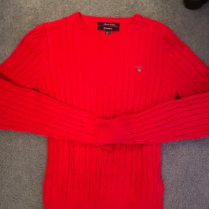 Röd kabelstickad tröja från GANT - Säljer en röd kabelstickad tröja från GANT i bomull. Tröjan har rund halsringning och långa ärmar. Klassisk och stilren design med diskret GANT-logo på bröstet. Perfekt för dig som gillar tidlös stil.