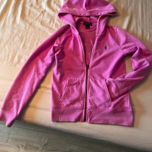 Rosa hoodie från Polo Ralph Lauren - Säljer en rosa hoodie från Polo Ralph Lauren med dragkedja och huva. Tröjan har långa ärmar, två fickor framtill och en liten blå logga broderad på bröstet. Perfekt för en avslappnad stil.