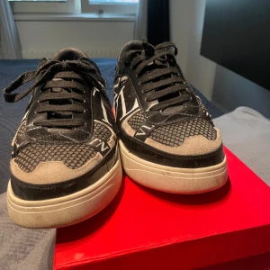 Svarta sneakers från Valentino Garavani - Säljer ett par svarta Valentino Garavani sneakers med vita detaljer och logga baktill. Skorna har snörning, rund tå och platt sula. Snyggt mönstrad ovandel i mocka och syntet. Kommer med originalkartong. 2 år gammla väldigt sällan använda men har använd dom ett par antal gånger!
