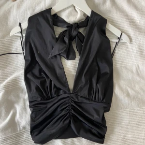 Svart omlottopp med knyt och rynkdetalj - Snygg svart omlottopp med djup v-ringning och knyt i nacken. Toppen har rynkdetalj framtill och är ärmlös, perfekt för en trendig look.