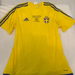Gul fotbollströja från Adidas med Nassjö-emblem - Säljer en gul kortärmad fotbollströja från Adidas med blå detaljer och Nassjö-emblem på bröstet. Tröjan har blå ränder på axlarna och tryck från matchen Sverige vs Luxemburg 2017. Perfekt för fotbollsfans!