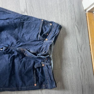 Mörkblå jeansbyxor med raka ben - Mörkblå jeansbyxor med klassisk femficksdesign och raka ben. Byxorna har kontrastsömmar och knappgylf. Modellen är i storlek S och har short length. 