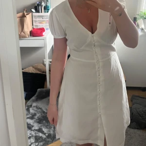 Vit kortklänning från Nelly  - Supersöt vit klänning från Nelly med korta puffärmar och v-ringning. Klänningen har dekorativa knappar framtill och en figurnära passform som markerar midjan. Perfekt för sommarens alla tillfällen! Aldrig använd. Hör av er vid fler frågor eller bilder!💛