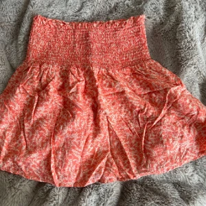 Korallröd blommig kortkjol från VAS - Supersöt korallröd kortkjol från YAS med vitt blommönster och smockad midja. Kjolen har en luftig och flowy passform som ger en avslappnad vibe. Perfekt att styla med en stickad tröja eller som en enkel topp.