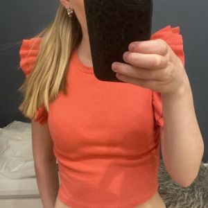 Koralrosa croppad topp med volangärmar - Säljer en korallrosa croppad topp med volangdetaljer på axlarna. Toppen har en tight passform och är perfekt för dig som vill sticka ut lite extra. Superfin att matcha med både jeans och kjol!