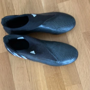 Svarta adidas predator fotbollsskor utan snörning - Säljer ett par svarta adidas fotbollsskor med blå och röda detaljer på sidan. Skorna har en strömlinjeformad design utan snörning och mönstrad ovansida. Perfekta för dig som vill ha bra grepp på planen. Ena mitten dobben på höger sko är lite nedgången. Annars väldigt sparsamt använda och som nya i övrigt. 