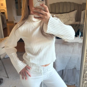 Vit ribbad croppad stickad tröja - Säljer en vit ribbad stickad tröja med snygg passform och långa ärmar. Tröjan har en hög hals och är mjuk och skön, perfekt för kyligare dagar. Enkel och stilren design som passar till mycket.