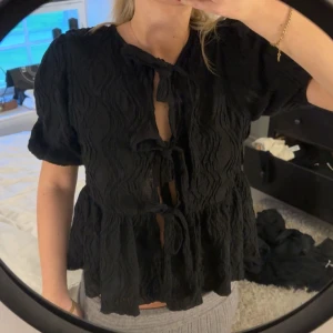 Svart blus med knyt fram - Svart blus i strukturerat tyg med korta puffärmar och tre knytningar framtill. Blusen har en lös passform och volang nedtill, vilket ger en trendig och avslappnad look.