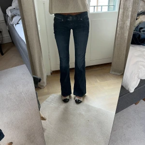 Mörkblå bootcut jeans från Guess - Snygga mörkblå jeans från Guess med bootcut passform. Jeansen har klassiska fem fickor, låg midja och detaljerad logotyp på bakfickan, storlek 34. Sitter snyggt och är i fint skick. Jag är 169 cm lång, och på bilderna är mitt innerbensmått ca 82,5 cm och midjemått (platt mätt på mig) ca 31 cm.