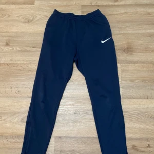 Svarta träningsbyxor från Nike - Säljer ett par svarta träningsbyxor från Nike med vit logga på ena låret. Byxorna har elastisk midja och smal passform, perfekta för sport eller gym. Materialet är syntetiskt och känns lätt och smidigt. Om ni har frågor då kan ni skicka till mig.