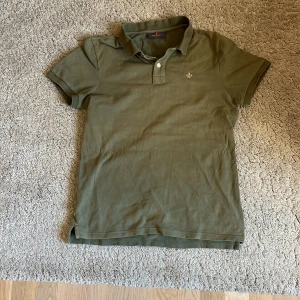 Olivgrön pikétröja från Morris - Snygg olivgrön pikétröja från Morris med klassisk krage och två knappar framtill. Tröjan har korta ärmar och en broderad logga på bröstet. Perfekt för en stilren och avslappnad look. 