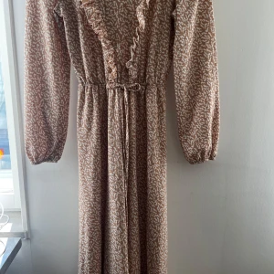 Beige långklänning - Långklänning i beige och vitt med abstrakt mönster. Klänningen har långa ärmar, volangdetaljer längs ringningen och knyt i midjan. Klänningen är från Modanisa. Någon söm har gått upp vid handleden på högersida. Men det är inte som påverkar funktionen och kan lagas med två stygn 