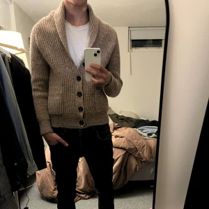 Beige stickad kofta från Ted Bernhardtz - Mysig beige stickad kofta från Ted Bernhardt med grov ribbstickning och stora svarta knappar framtill. Koftan har två fickor och en bred sjalkrage. Perfekt för lager-på-lager och en avslappnad stil.