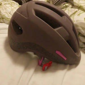 Lila cykelhjälm med rosa detaljer - Säljer en lila cykelhjälm med rosa och röda detaljer. Hjälmen har flera ventilationshål och justerbart spänne under hakan. Perfekt för dig som vill ha en snygg och säker hjälm till cykelturen.