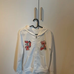 Vit hoodie med dragkedja och tryck - Vit hoodie med dragkedja framtill och huva. På framsidan finns ett tryck med bokstaven P och en nalle med bokstaven R i rött och svart. Tröjan har långa ärmar och två fickor framtill. Tröjan är väldigt tunn men perfekt för en sommardag!