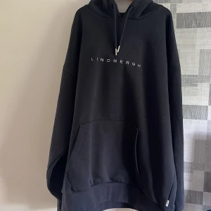 Svart oversized hoodie från Lindbergh - Säljer en svart oversized hoodie från Lindbergh med vit text på bröstet. Tröjan har huva med dragsko och en stor magficka framtill. Perfekt för en avslappnad stil.