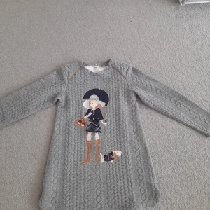 Grå stickad klänning med broderad figur - Säljer en grå stickad klänning med lång ärm och ett broderat motiv av en tjej med hund framtill. Klänningen har ett kabelstickat mönster och är perfekt för dig som gillar unika detaljer.