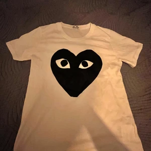 Vit t-shirt med svart hjärta och ögon - Vit t-shirt från PLAY med ett stort svart hjärta med ögon tryckt på bröstet. Klassisk passform och rund halsringning. Perfekt för dig som gillar grafiska tryck och vill sticka ut lite extra.